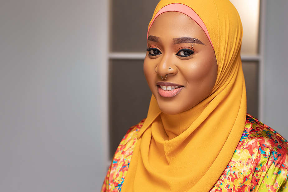 Portrait of Hauwa Ojeifo, who wears a yellow and pink hijab headscarf and has silver piercings on her left eyebrow and nose. © Hauwa Ojeifo