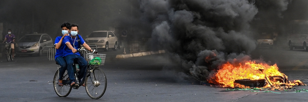 Une photo prise le 3 avril 2021 montre des personnes passant à vélo devant une barricade de fortune en feu, érigée par des manifestants contre le coup d’État militaire, dans la commune de Tamwe, à Yangon. © AFP