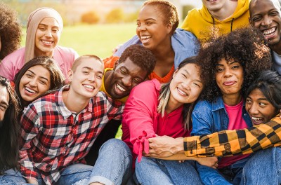 A group of people of diverse ethnicities sit together, hug, and laugh. © Getty Images/DisobeyArt