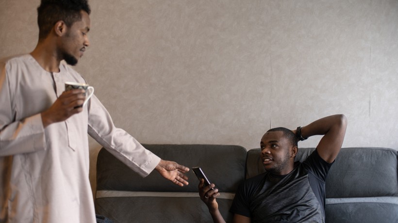 A man standing with his arm outstretched towards a phone held by a man sitting on a sofa