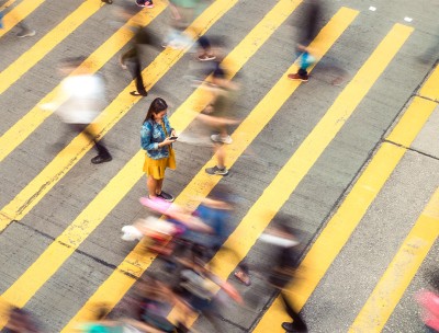 A woman pausing to focus on a message on her phone while on a busy crosswalk, with motion blur as people move around her. © Getty Images