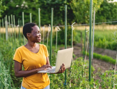 A woman holding a laptop looks at a farming field while she smiles. © ijubaphoto / Getty Images