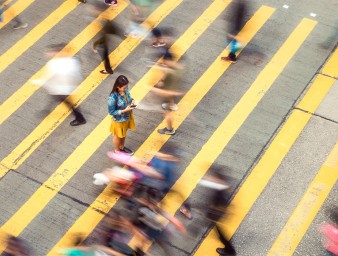 A woman pausing to focus on a message on her phone while on a busy crosswalk, with motion blur as people move around her. © Getty Images