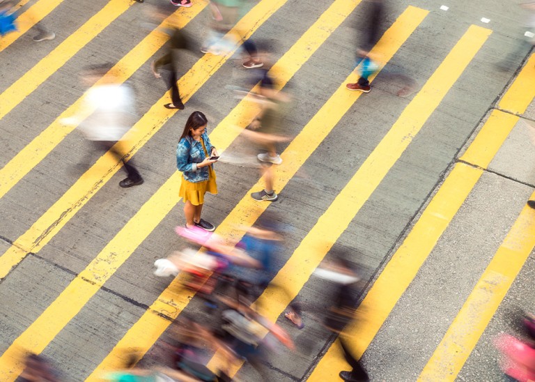 A woman pausing to focus on a message on her phone while on a busy crosswalk, with motion blur as people move around her. © Getty Images